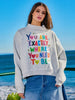 Brunette woman wearing a gray hoodie with colorful text  "You are exactly where you need to be" by the ocean