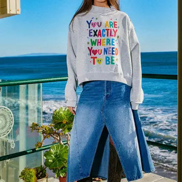 Brunette woman wearing a sweatshirt with motivational text "You are exactly where you need to be" by the ocean